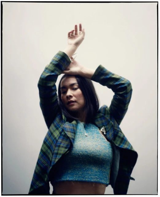Portrait of a young woman with her arms raised, wearing a blue and green plaid jacket and blue shirt, against a light grey background.