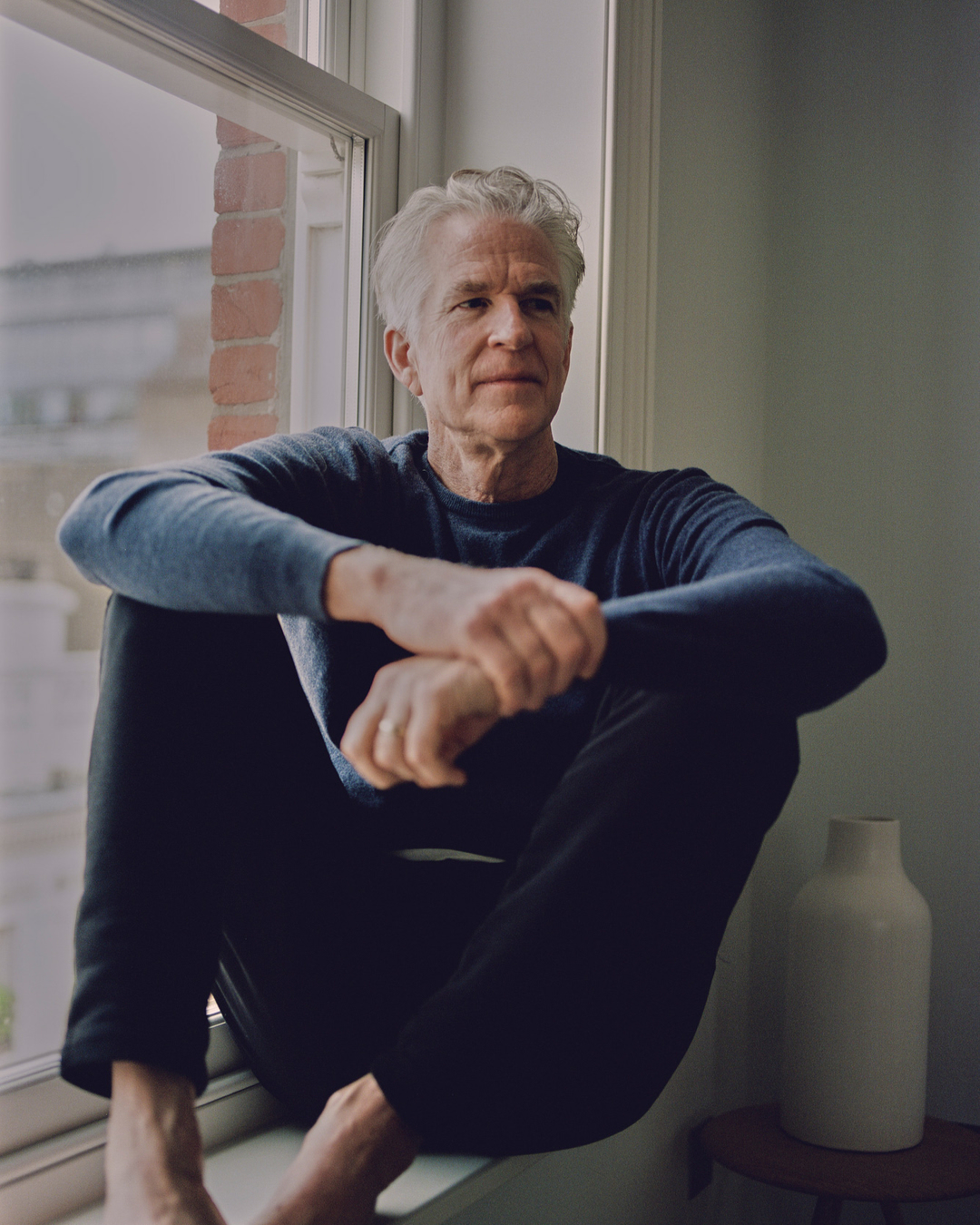 Mature man in blue sweater sits cross-legged in front of a bright window in an indoor setting. Natural light illuminates the scene.