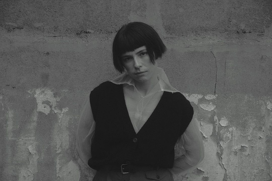 Monochromatic portrait of a fair-skinned adult female with dark blunt bangs wearing a black vest and sheer white blouse indoors.