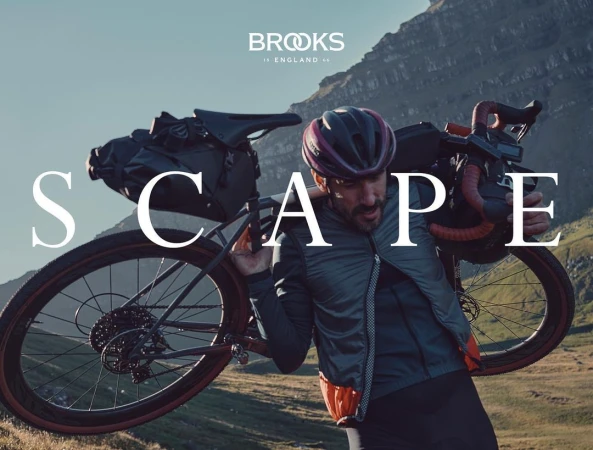 Man carries bike on shoulder in outdoor setting with mountain backdrop; Brooks England logo, emphasizing sport, nature, and product.