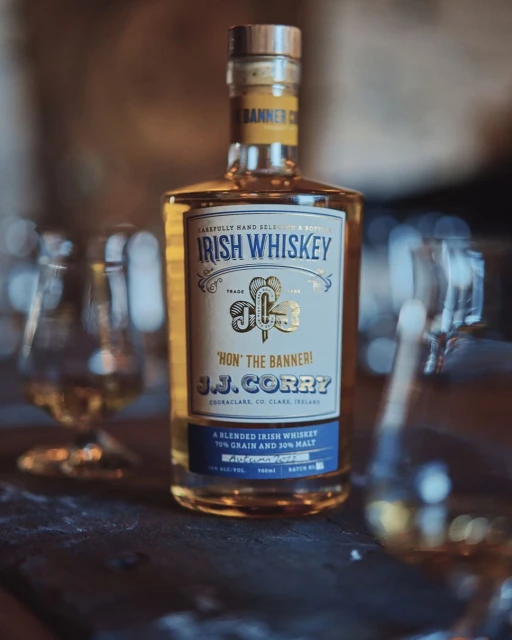 Bottle of J.J. Corry Irish Whiskey on a dark wooden surface with blurred glassware in the background, advertising the liquor.