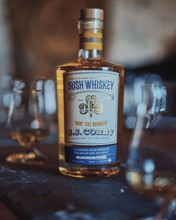 Bottle of J.J. Corry Irish Whiskey on a dark wooden surface with blurred glassware in the background, advertising the liquor.