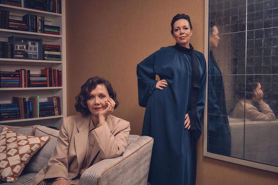 Two women pose indoors; one is seated on a patterned chair with a bookshelf behind her, the other stands next to a mirror.