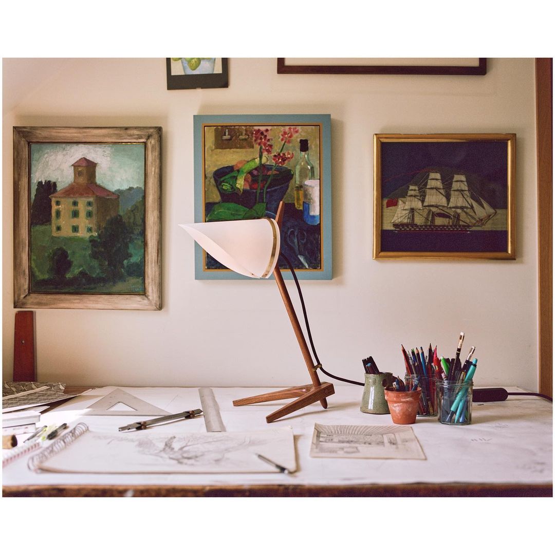 Interior shot of a desk with a drafting lamp, drawing tools, and framed paintings in the background, creating a creative workspace.