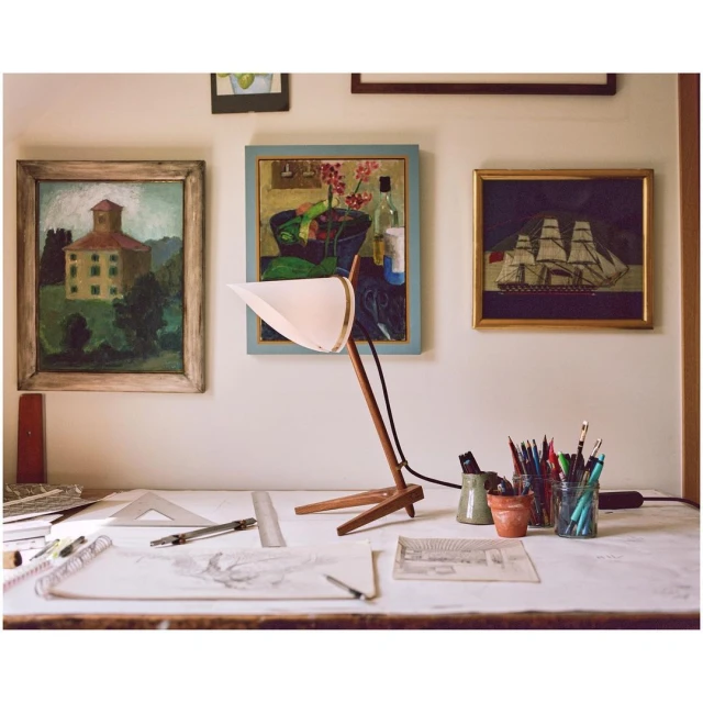 Interior shot of a desk with a drafting lamp, drawing tools, and framed paintings in the background, creating a creative workspace.