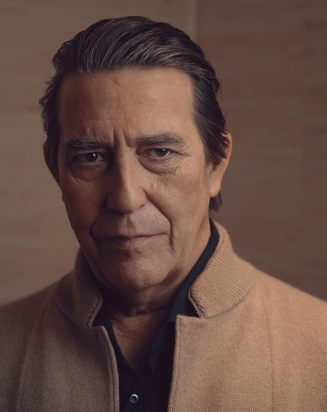 Portrait of an adult man with dark hair wearing a tan jacket and dark shirt in an indoor setting with neutral background.