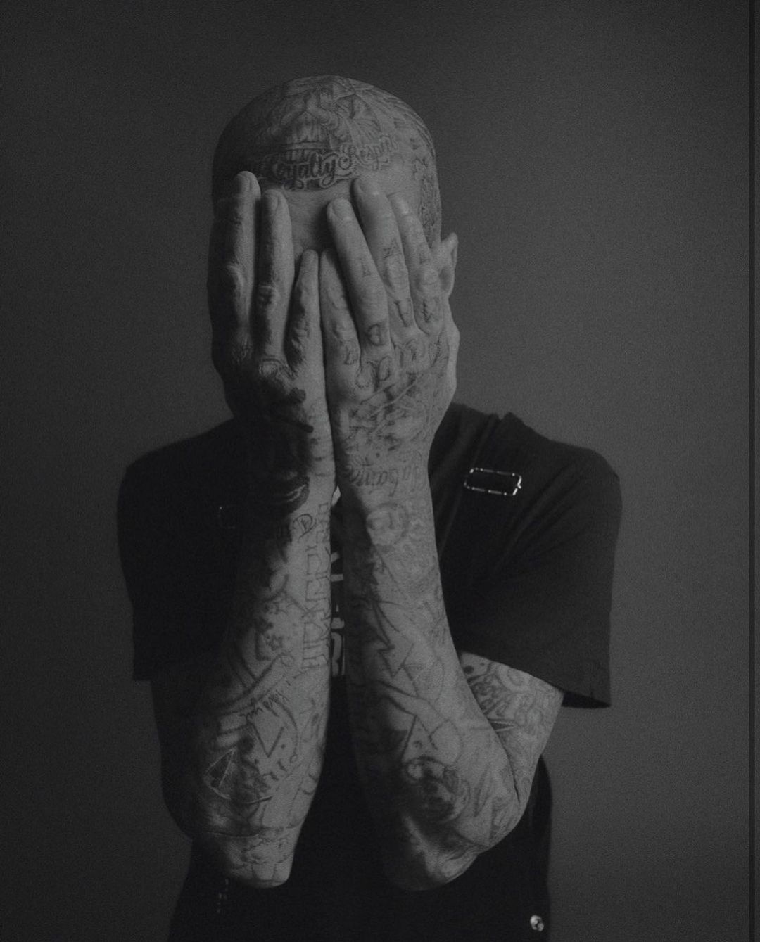 Monochrome portrait of a tattooed adult with hands covering face against a grey backdrop, showcasing detailed body art and minimal lighting.