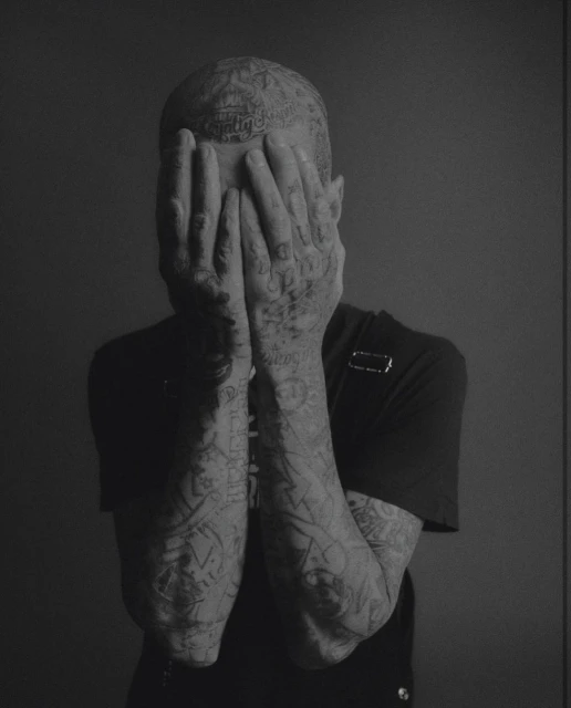 Monochrome portrait of a tattooed adult with hands covering face against a grey backdrop, showcasing detailed body art and minimal lighting.