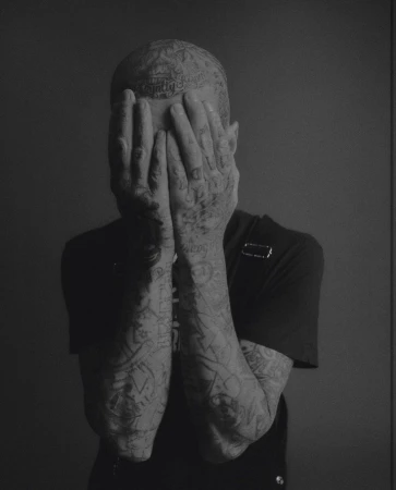 Monochrome portrait of a tattooed adult with hands covering face against a grey backdrop, showcasing detailed body art and minimal lighting.