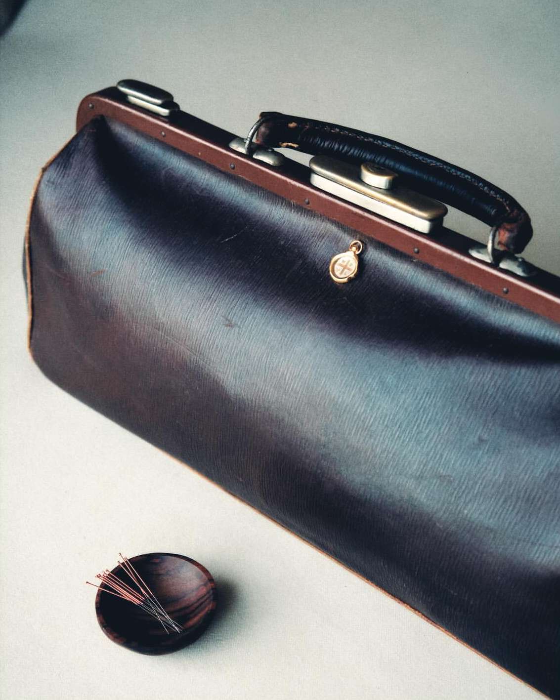 A brown leather doctor's bag with metal accents and a small dish of acupuncture needles sits on a light surface.
