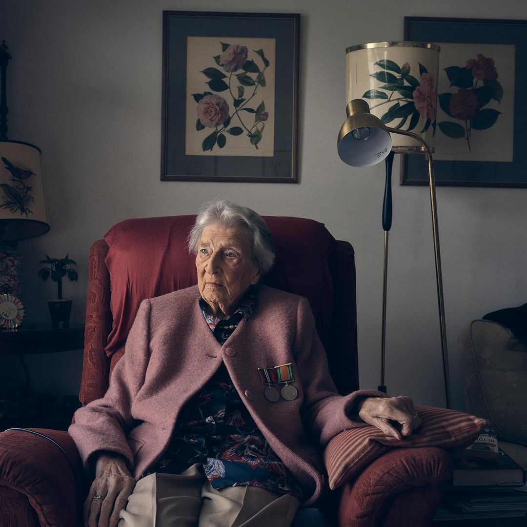 Elderly woman in a red armchair with floral paintings on the wall and a brass floor lamp, creating a calm indoor scene.
