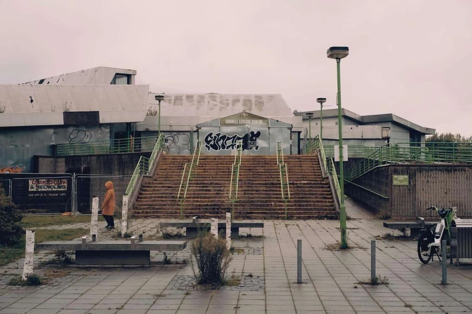 Exterior view of a concrete building with stairs leading up to the entrance, a person in a yellow jacket walks near benches.