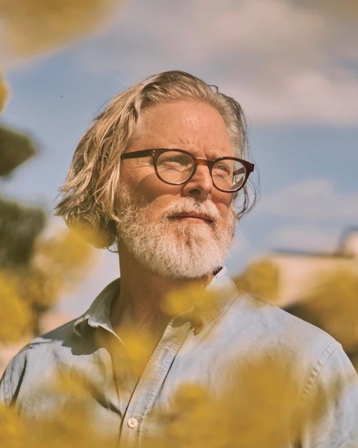 Portrait of an adult male with long blonde hair, a white beard, and glasses, wearing a light blue shirt outdoors on a sunny day.