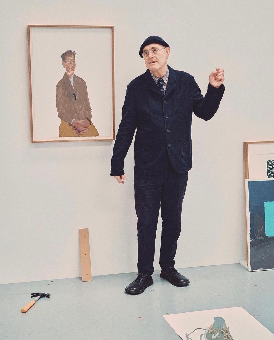 An adult male stands in an art studio next to a framed painting of a seated male, with a hammer, wood plank, and other art nearby.