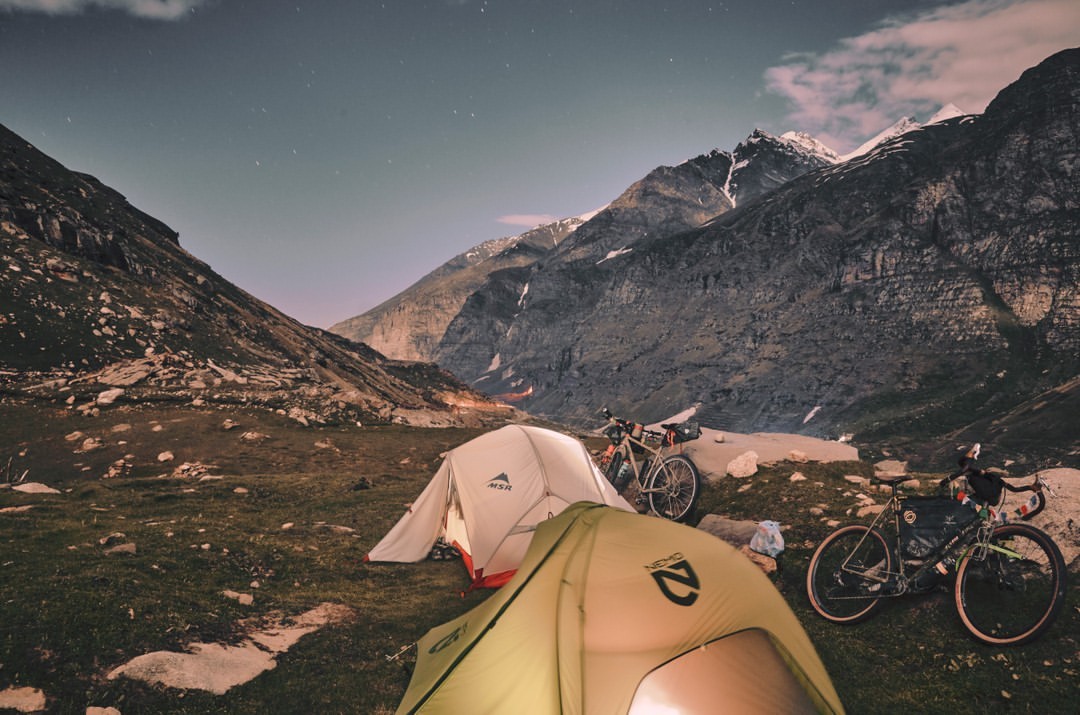 Camping scene at night featuring two tents, mountain bikes, and a backdrop of mountainous terrain under a starlit sky.