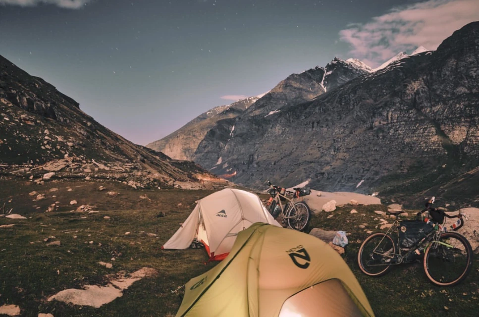 Camping scene at night featuring two tents, mountain bikes, and a backdrop of mountainous terrain under a starlit sky.