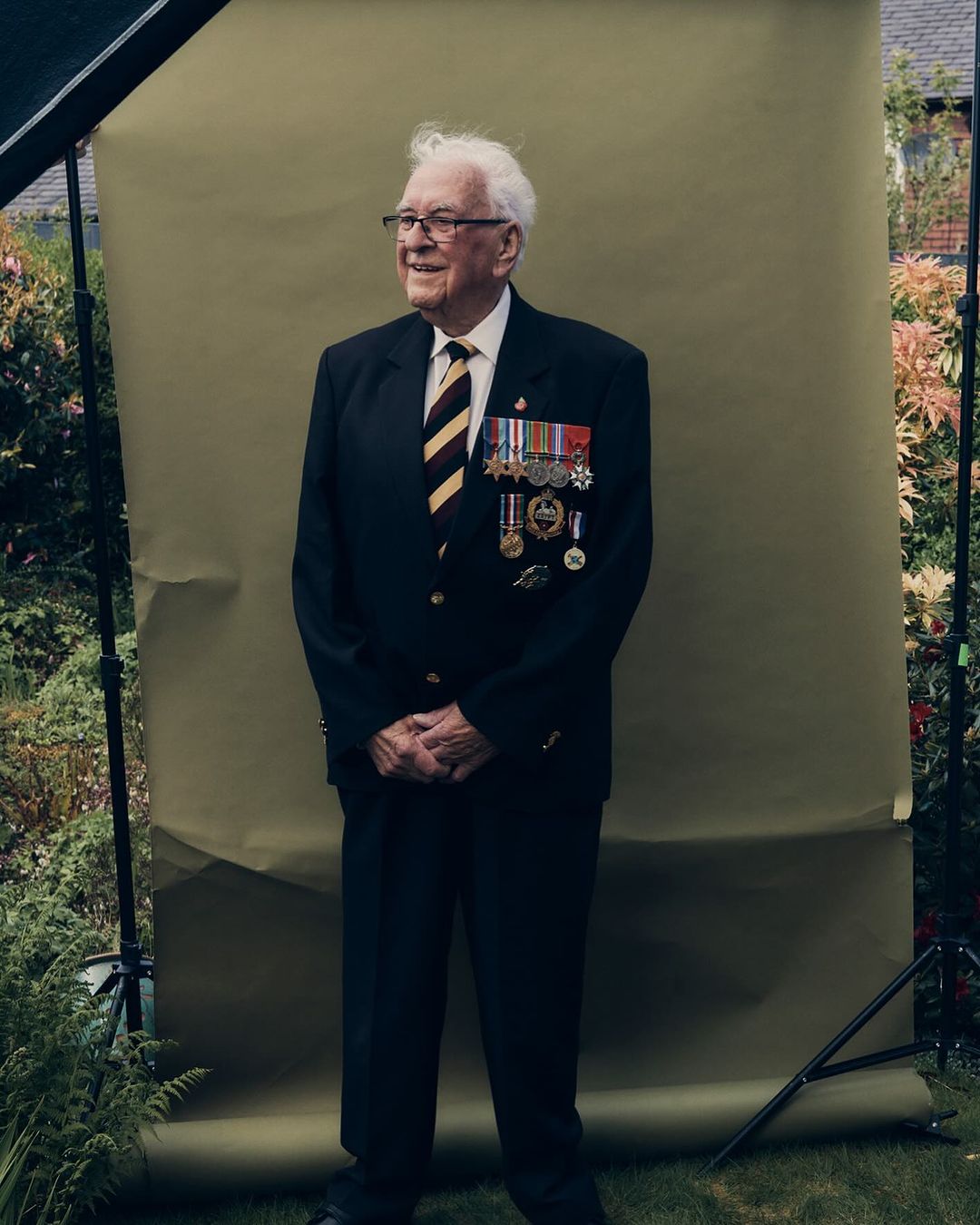 Elderly man in suit and tie stands in front of a green backdrop in garden; wearing medals and glasses, he is smiling slightly.