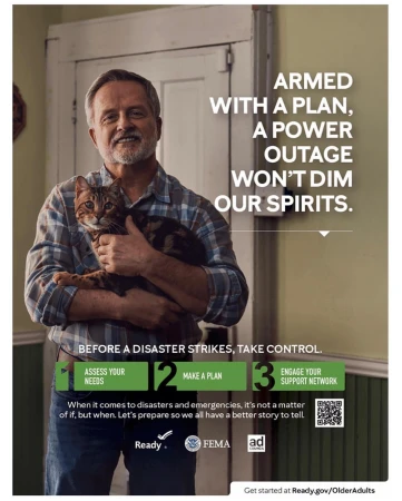 Middle-aged man in a plaid shirt smiles while holding a cat, next to a poster promoting disaster preparedness, indoor setting.