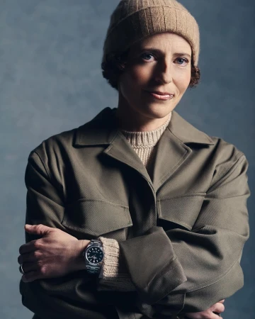 Portrait of a woman wearing an olive green jacket, knit beanie, and metal wristwatch with arms crossed