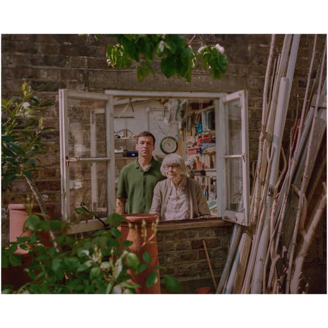 An adult male and elderly woman pose by an open window on a brick wall, surrounded by plant pots, plants, and wooden beams.