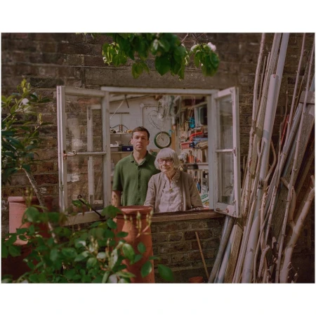 An adult male and elderly woman pose by an open window on a brick wall, surrounded by plant pots, plants, and wooden beams.