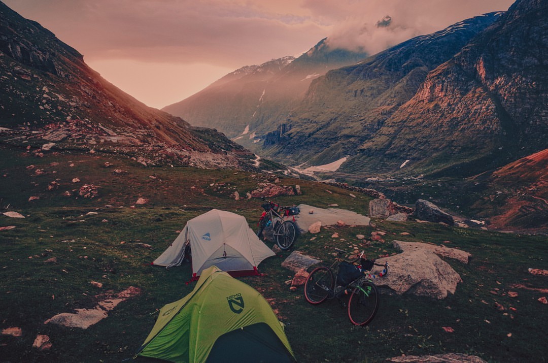 Two tents and two bicycles are set up in a mountainous landscape under a dramatic sky, creating a scene of outdoor adventure.