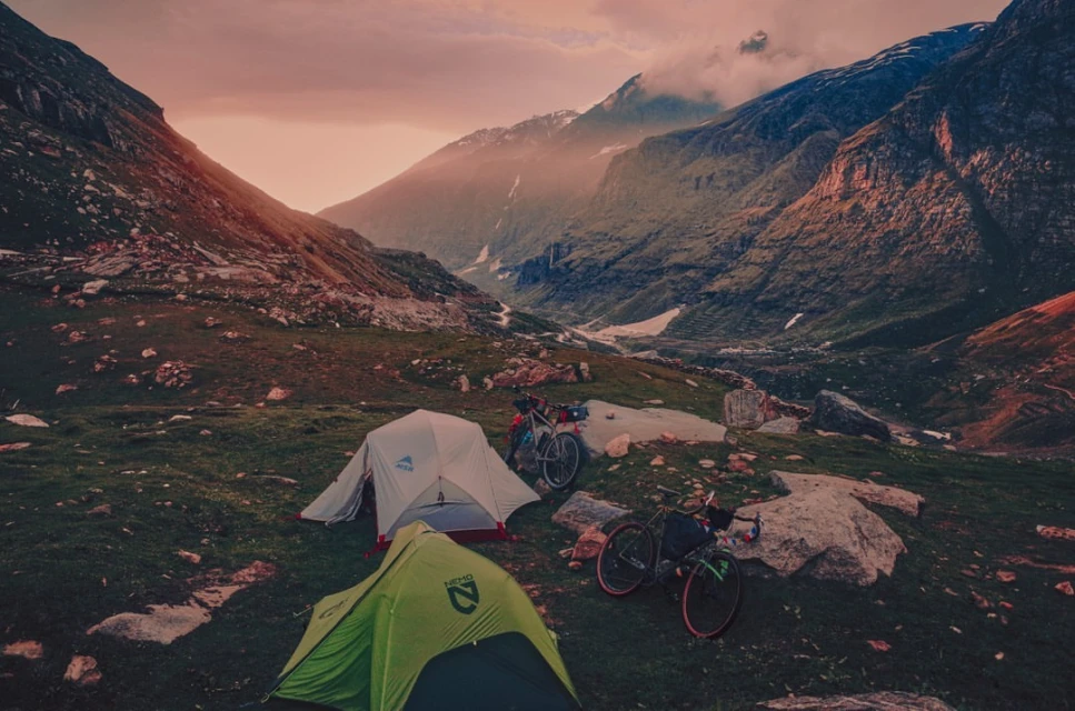 Two tents and two bicycles are set up in a mountainous landscape under a dramatic sky, creating a scene of outdoor adventure.