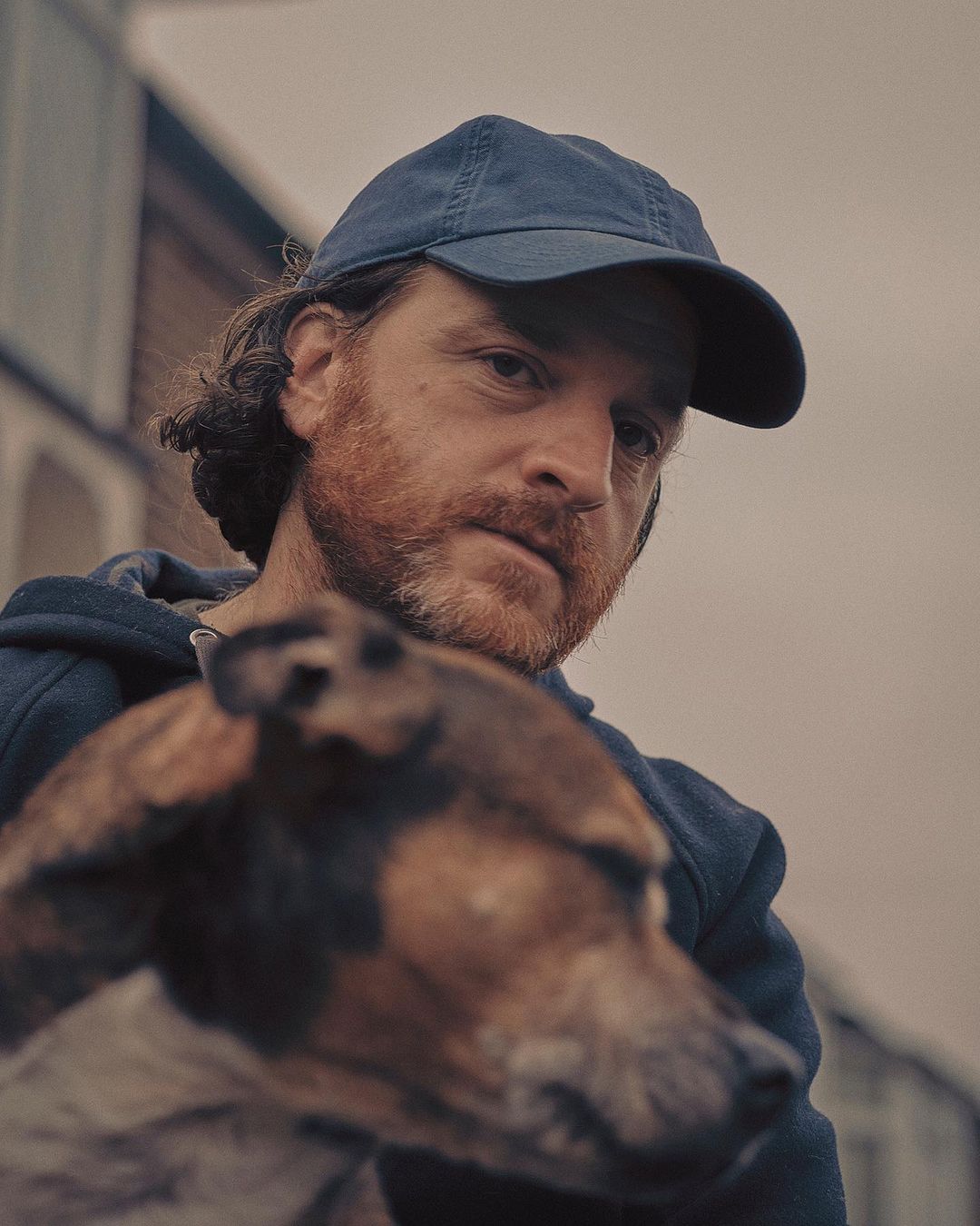 Man wearing a blue baseball cap and dark hoodie holding a small brown dog outdoors under an overcast sky.