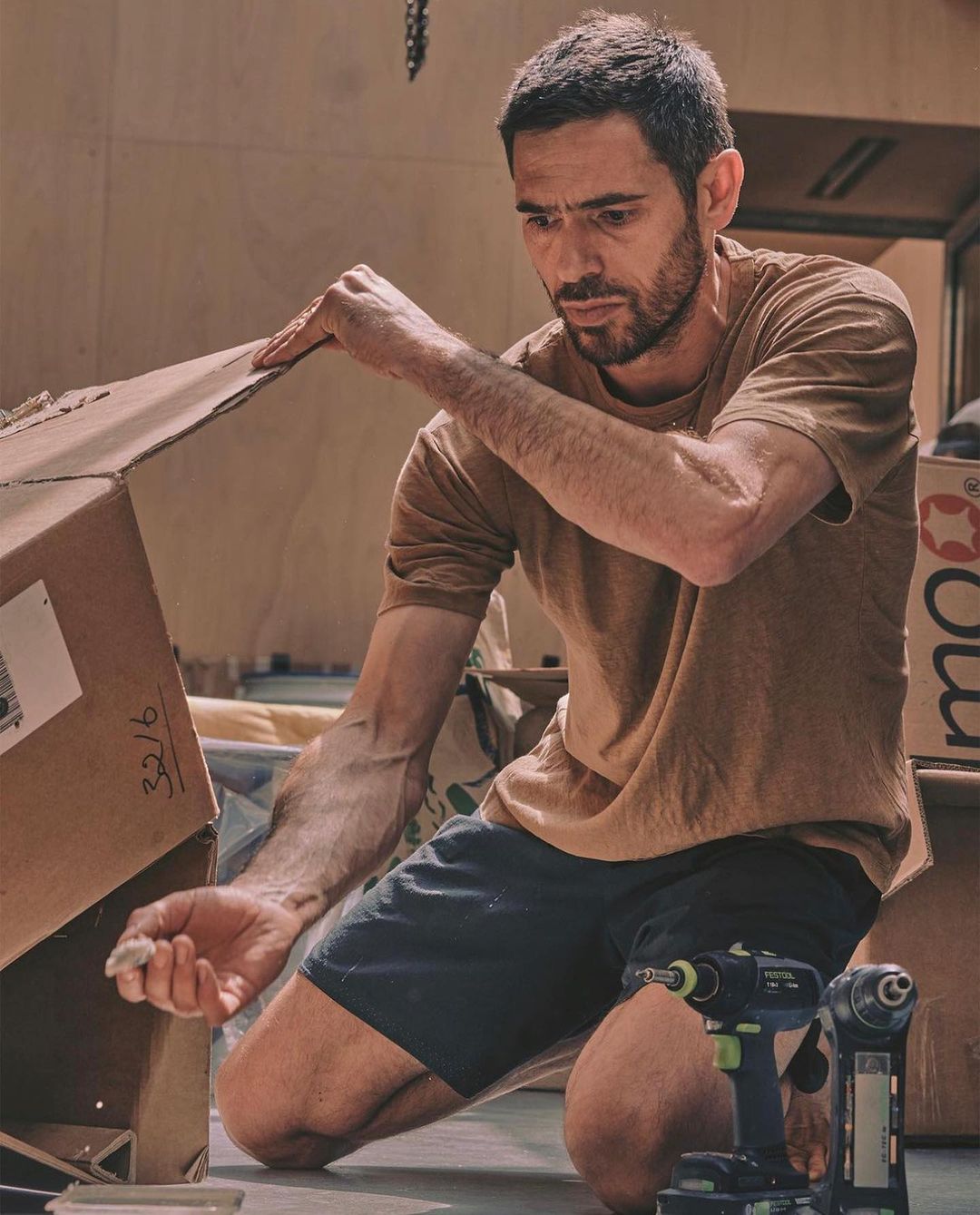 Adult male kneeling indoors assembling a cardboard box with hand tools nearby in warm, diffused lighting and shallow depth of field.