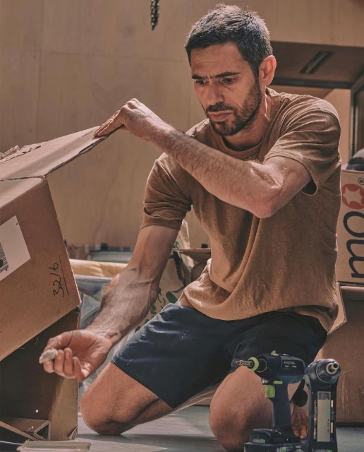 Adult male kneeling indoors assembling a cardboard box with hand tools nearby in warm, diffused lighting and shallow depth of field.