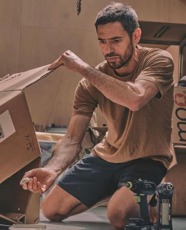 Adult male kneeling indoors assembling a cardboard box with hand tools nearby in warm, diffused lighting and shallow depth of field.