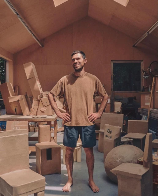 Smiling man in a brown shirt stands in a workshop filled with cardboard furniture. Natural light from skylight and window.