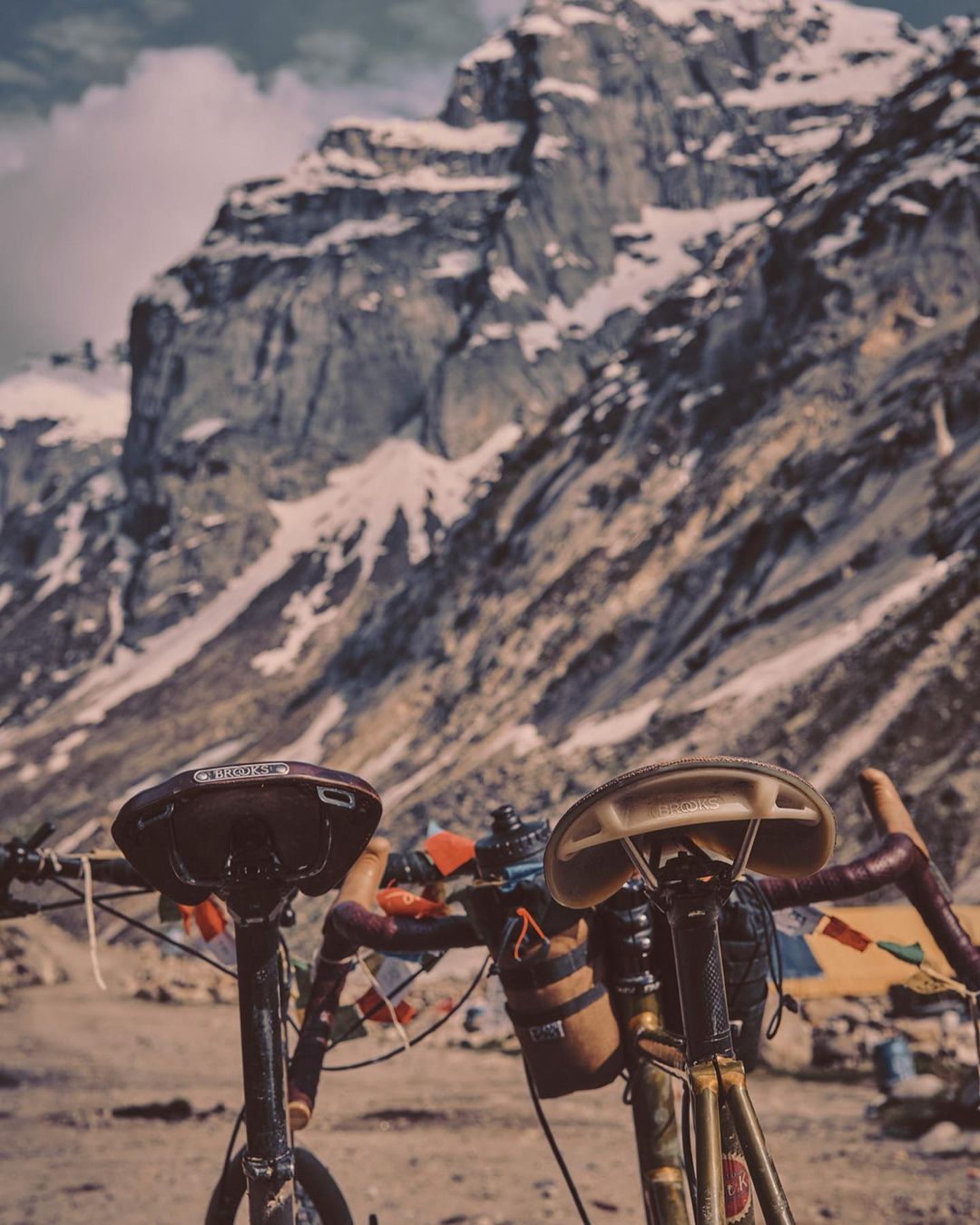 Two touring bicycles with accessories sit on a dirt ground with a backdrop of massive mountains and clouds. Cloudy skies above.