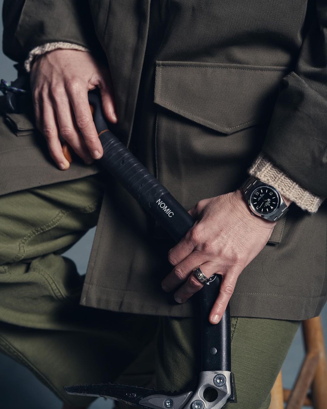 Person holding ice axe, wearing olive green jacket, cargo pants, and a wristwatch; showcasing outdoor gear and accessories in a studio setting.