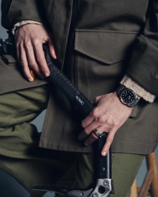 Person holding ice axe, wearing olive green jacket, cargo pants, and a wristwatch; showcasing outdoor gear and accessories in a studio setting.