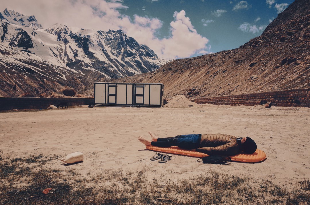 Adult male rests on an orange mat in a mountainous landscape with a white modular building, rocks, and a partly cloudy sky.