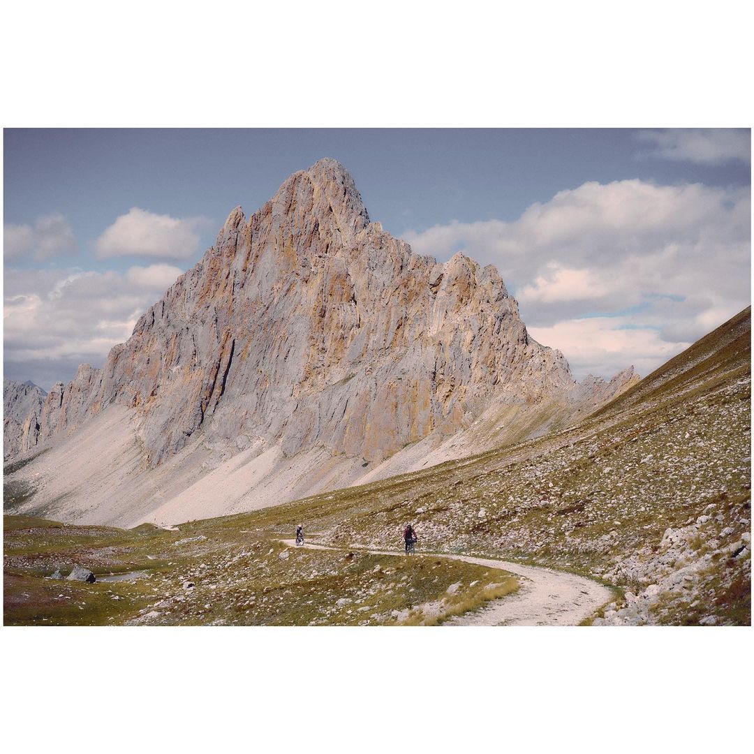 Scenic mountain landscape with a winding dirt path, two hikers, clear sky, and rocky mountain peak in the background.