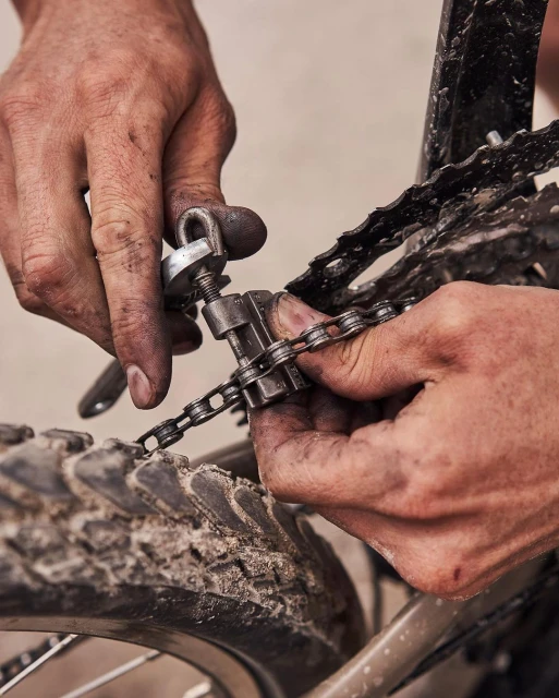 Close-up of person's hands using a chain tool to repair a bicycle chain on a muddy mountain bike tire with a neutral background.