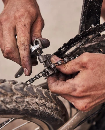 Close-up of person's hands using a chain tool to repair a bicycle chain on a muddy mountain bike tire with a neutral background.