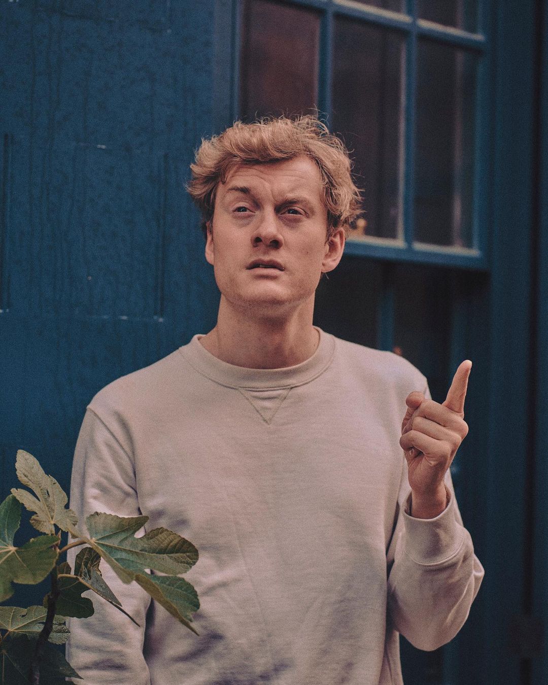 A young adult man with light hair poses in front of a blue building, pointing up with his right hand in a medium portrait shot.