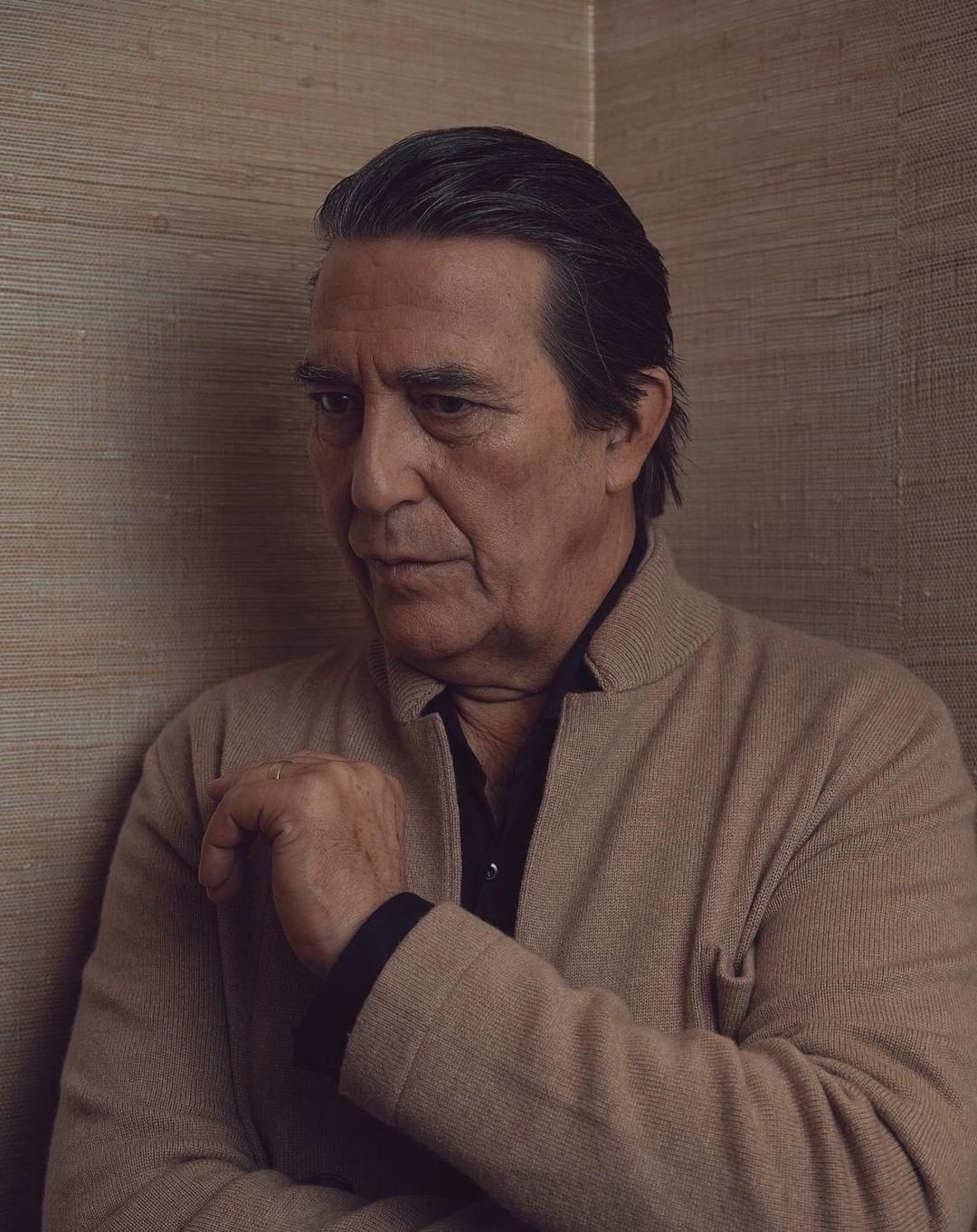 Portrait of a mature adult male with dark hair in a beige jacket against a textured wall, looking contemplative in soft, neutral lighting.