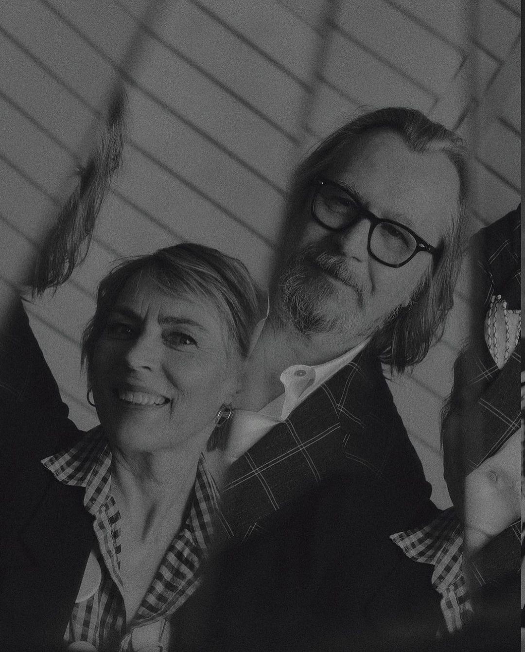 Monochrome portrait of a smiling woman and a man with a beard and glasses, posing indoors against a wall with diagonal stripes.