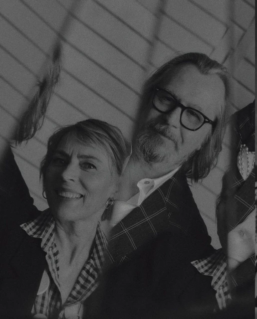 Monochrome portrait of a smiling woman and a man with a beard and glasses, posing indoors against a wall with diagonal stripes.
