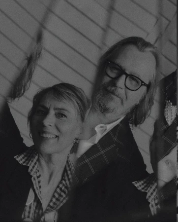 Monochrome portrait of a smiling woman and a man with a beard and glasses, posing indoors against a wall with diagonal stripes.