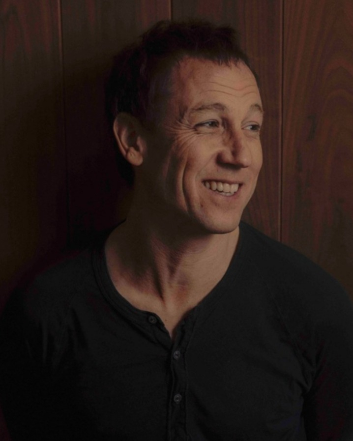 Portrait of a smiling man in a dark henley shirt standing against a warm wooden paneled wall