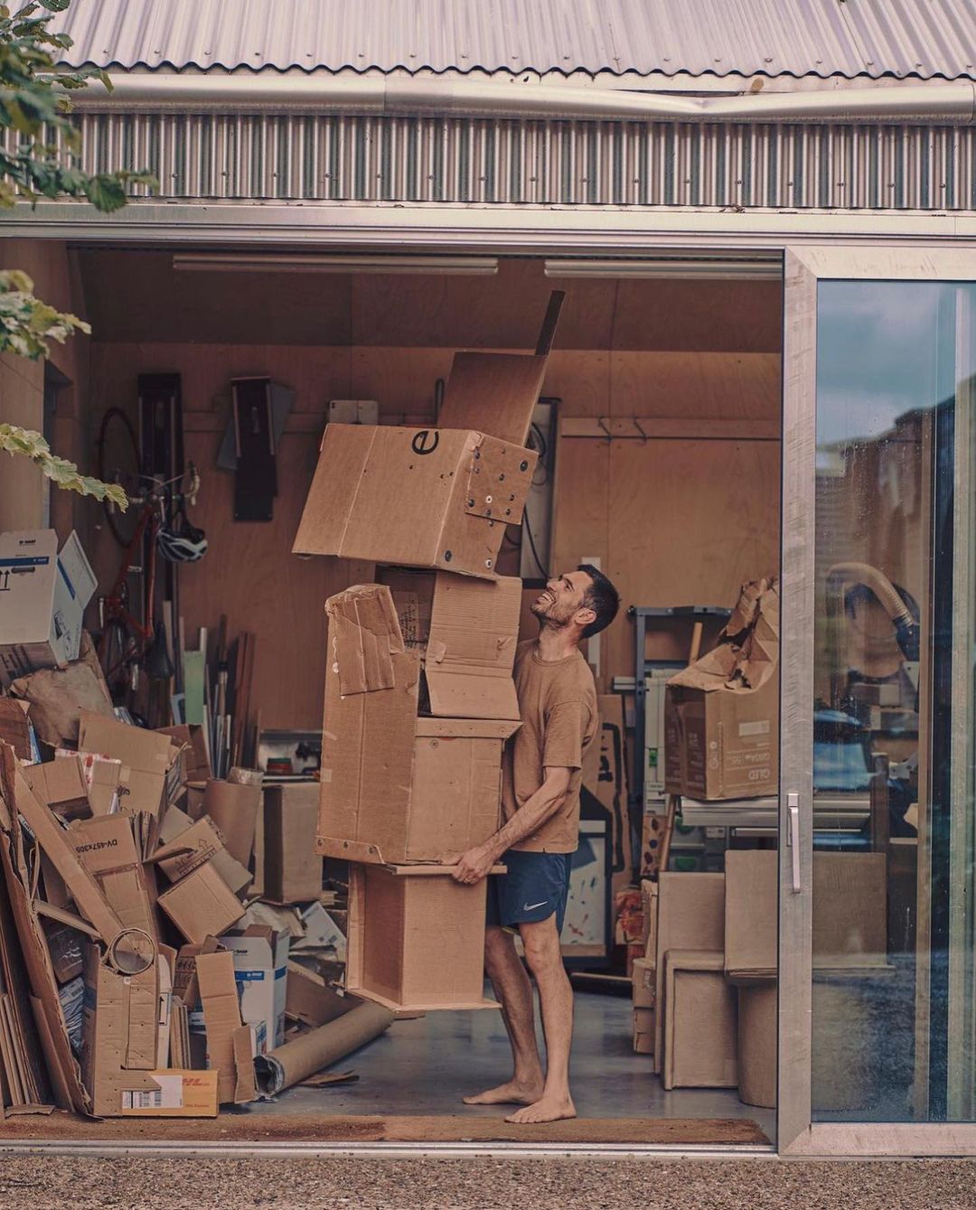 Adult male balancing a tall stack of cardboard boxes inside of a garage with additional cardboard boxes and a bicycle in the background.