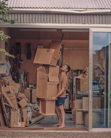 Adult male balancing a tall stack of cardboard boxes inside of a garage with additional cardboard boxes and a bicycle in the background.
