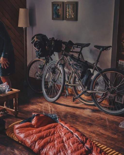 Indoor shot showcases two bicycles propped against a wall, with a red sleeping bag on the wooden floor, and warm lamp lighting.
