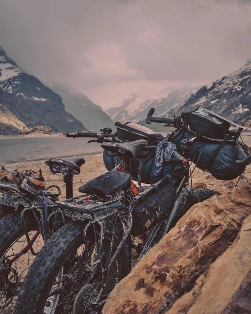 Two touring bikes loaded with gear are parked on a rocky lakeshore with snow-capped mountains in the distance under an overcast sky.