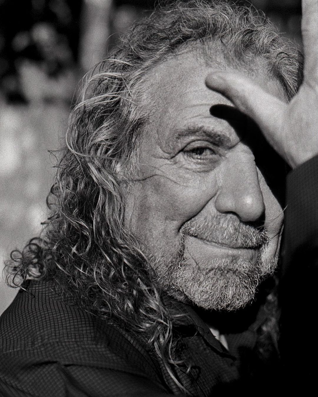 Black and white portrait of mature man with long curly hair, hand shielding face, wearing a collared shirt.